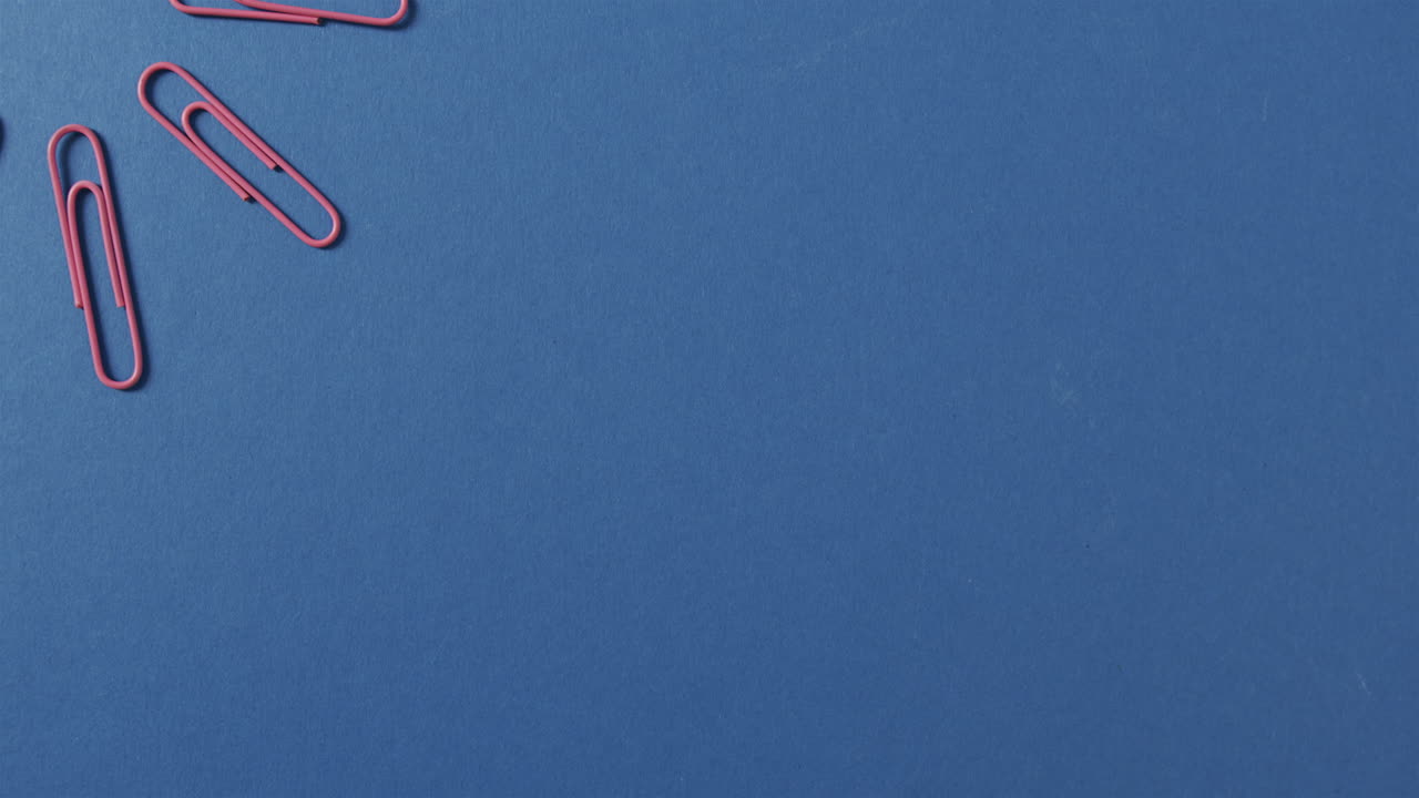 Vista dall'alto di graffette rosa disposte con spazio per il testo su sfondo blu, al rallentatore.
