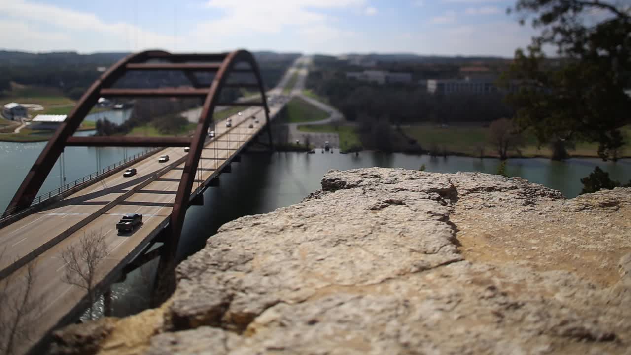 Austin Pennybacker Bridge tilt-shifted time-lapse, focus isolates section of ledge and front part of the bridge. Truck parked on boat ramp launches boat into the water; smoke seen as boat starts.