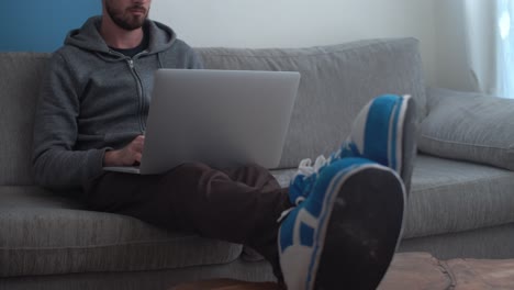 man sits comfortably on couch and works on laptop from home