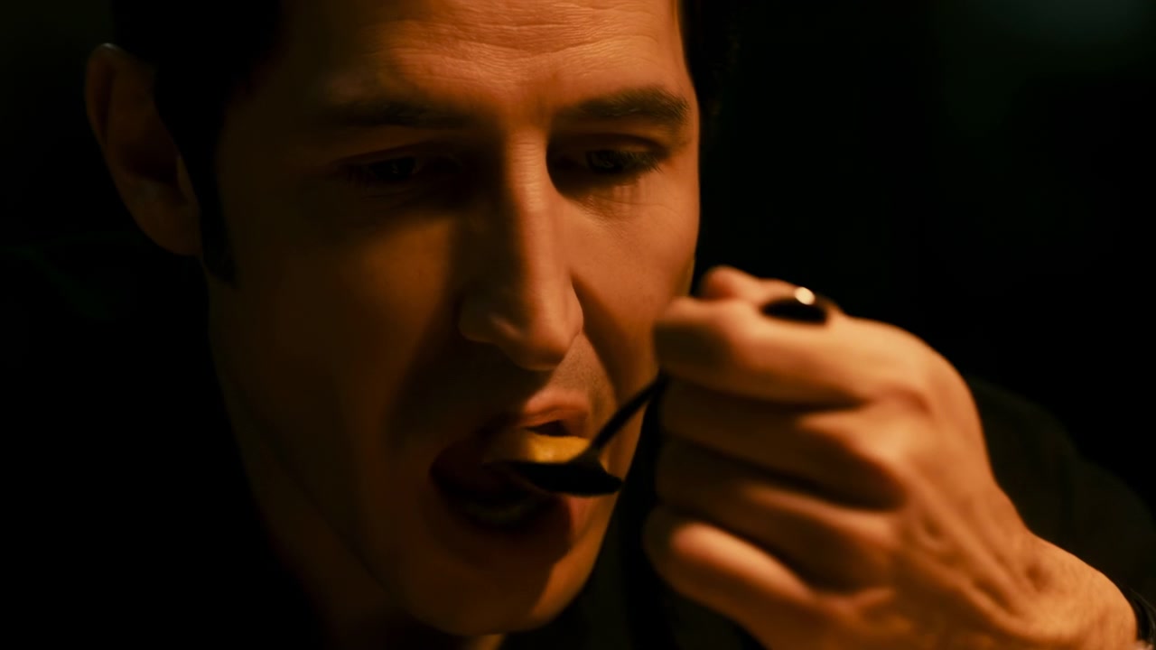 Un primo piano caldo e a bassa luminosità segue un uomo che guarda il cucchiaio, portando il cibo alle labbra, mangiando, poi masticando mentre la sua mano si abbassa, con ombre morbide e dintorni scuri.