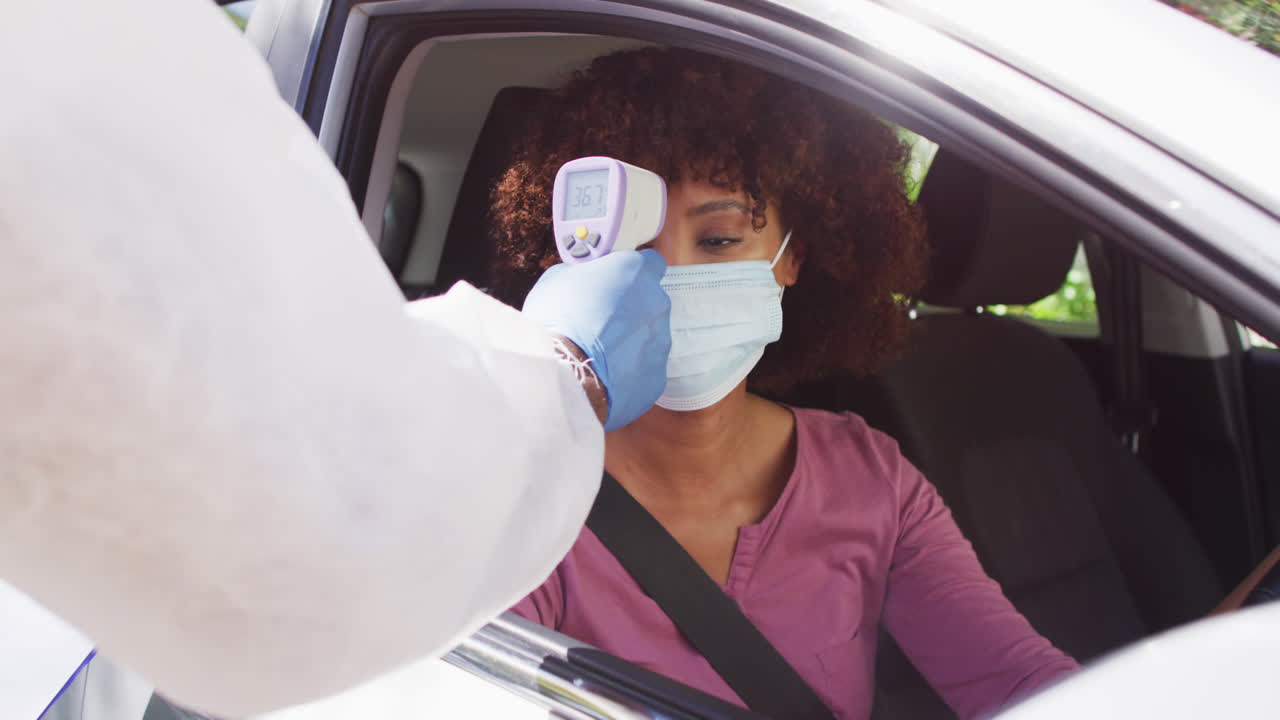 mujer afroamericana con máscara facial sentada en un coche con la temperatura medida por un trabajador médico