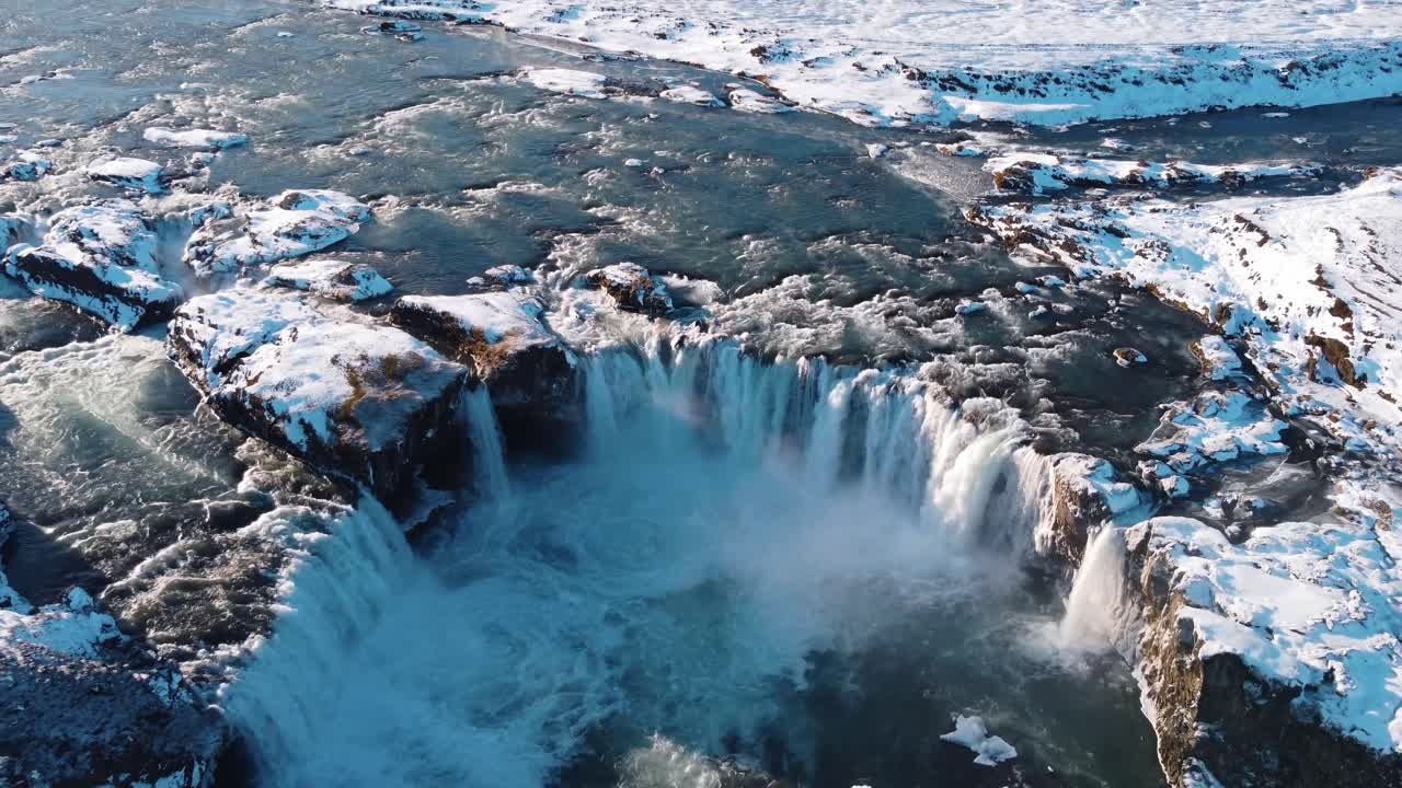 A dramatic aerial perspective showcases the powerful and iconic Godafoss waterfall, surrounded by snow and ice in the depths of winter