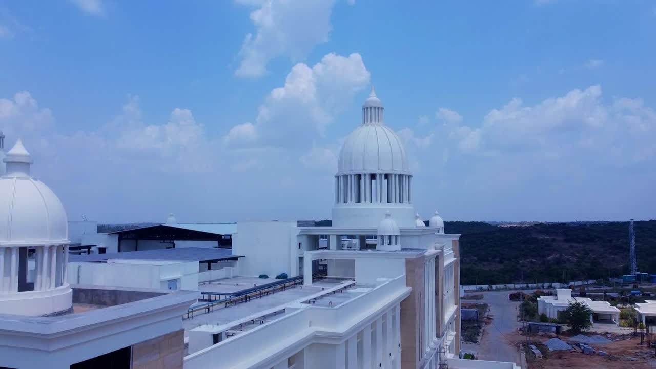 A smooth crane-up shot along the facade of the grand, neo-classical Kanha Shanti Vanam building in India, revealing its impressive scale and white domes against a beautiful blue sky with clouds