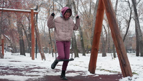 una mujer joven vestida con vestidos cálidos de invierno coloca la rodilla en un columpio de hierro en un parque al aire libre nevado, el suelo cubierto de nieve y los árboles desnudos crean una atmósfera fría mientras se equilibra en el columpio