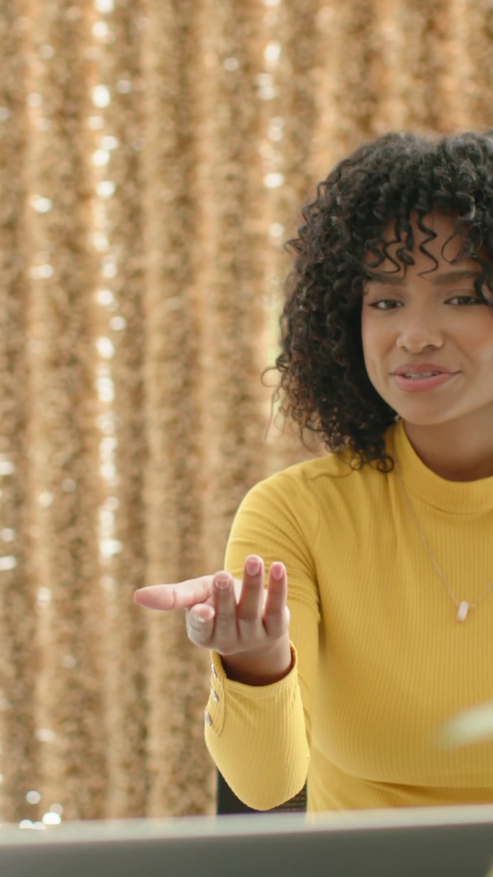 Vertical video: Smiling woman in gold top pointing at left hand over laptop and plant to discuss f
