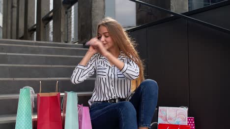 girl sitting on stairs with bags talking on mobile phone about sale in shopping mall in black friday