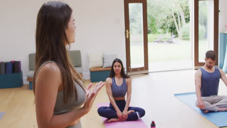 caucasian female instructor explaining to diverse man and woman sitting on mats at yoga class