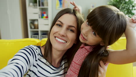 vista de cerca de la feliz madre e hija sentadas en un sofá amarillo, divirtiéndose, dándose afecto y mirando la cámara