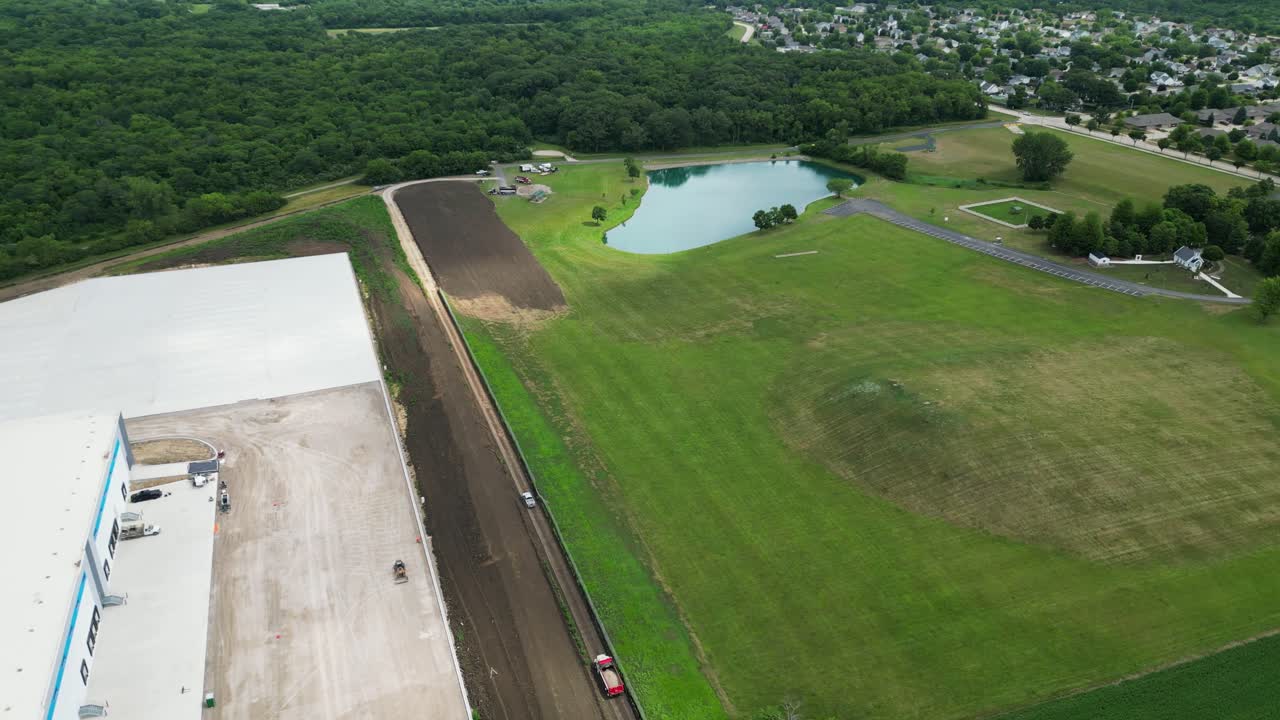 An aerial drone shot captures the scale of a new warehouse construction project bordering a lush green park area with a pond and nearby residential neighborhood