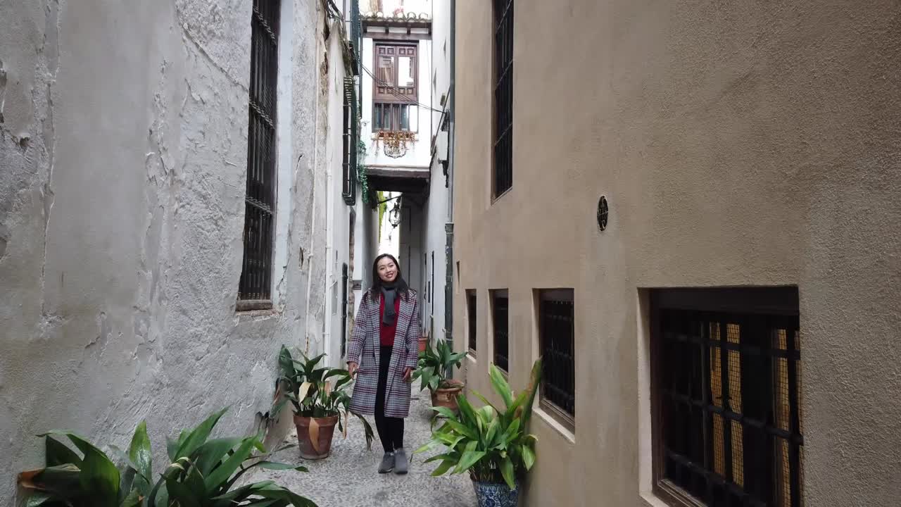 Young Asian woman approaches camera in backward tracking shot through Granada's narrow alley. White walls, wooden balconies, plants, lanterns. Dynamic urban travel footage