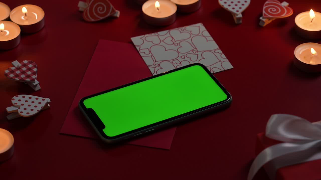 vista superior del teléfono inteligente con pantalla verde llave de croma en la mesa roja con velas encendidas, corazones, valentín y regalo. fondo crepúsculo romántico con luces. día de valentín. cerrar. cámara lenta