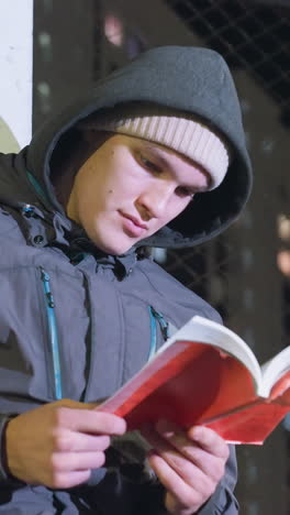 primer plano de un joven con capucha y gorra leyendo un libro de portada roja al aire libre por la noche, iluminado por las luces de la ciudad urbana con una valla de malla en el fondo