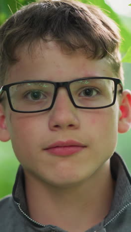 primer plano de un joven guapo con labios rosados, con gafas y una camiseta gris, mirando enfocado directamente a la cámara, está rodeado de vegetación con árboles borrosos en el fondo