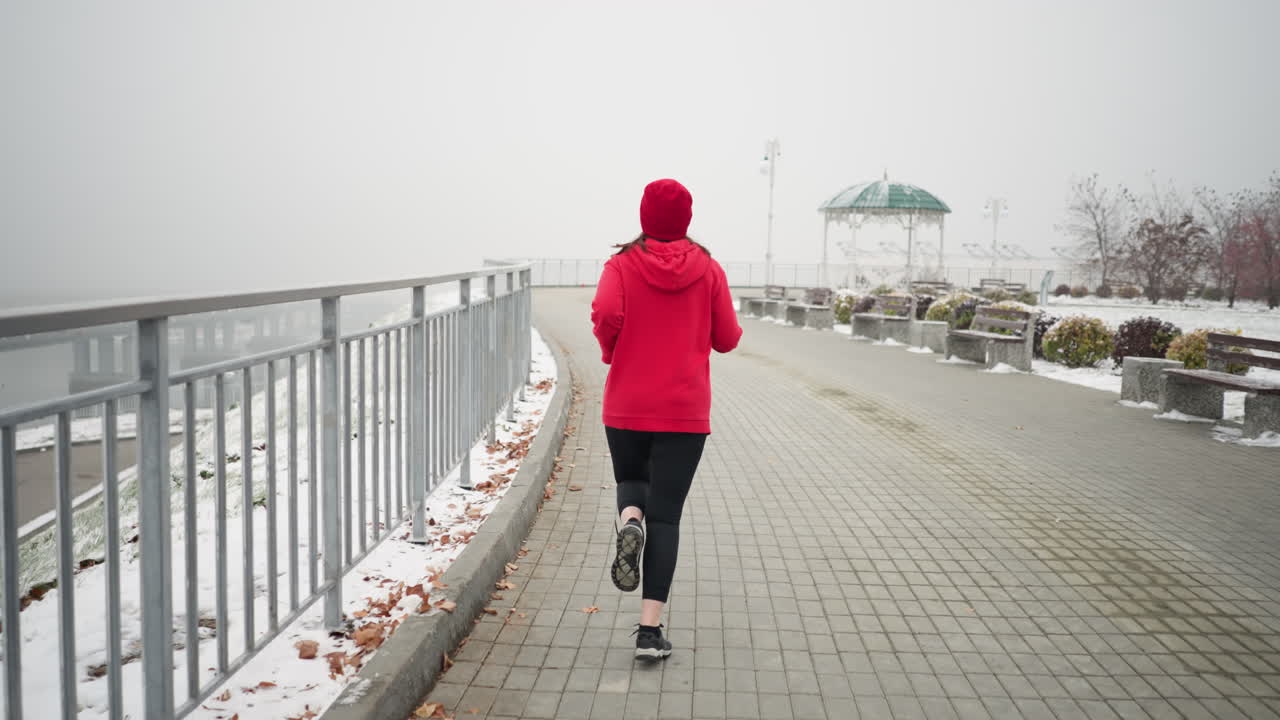Back view of lady jogging in red beanie and jacket along snowy pathway near iron railing, serene park setting, benches nearby, foggy atmosphere with distant bridge and winter trees