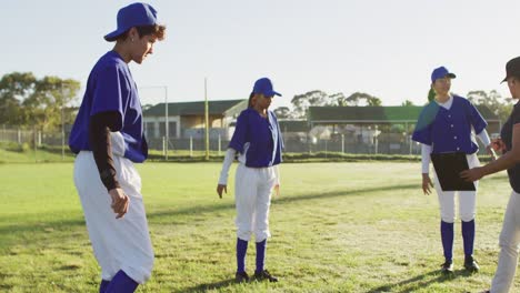 Diverse-group-of-female-baseball-players-with-coach,-warming-up-on-pitch,-standing,-stretching-legs