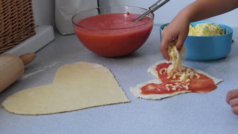 Die-Hand-Eines-Kleinen-Jungen-Mädchens-Streut-Mozzarella-Käse-über-Eine-Herzförmige-Pizza-In-Einer-Modernen-Küche,-Konzept-Für-Den-Familientag-Eines-Liebespaares