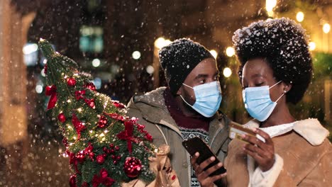 close-up view of joyful african american couple wearing facial masks talking and shopping online with smartphone while it¬¥s snowing on the street in christmas