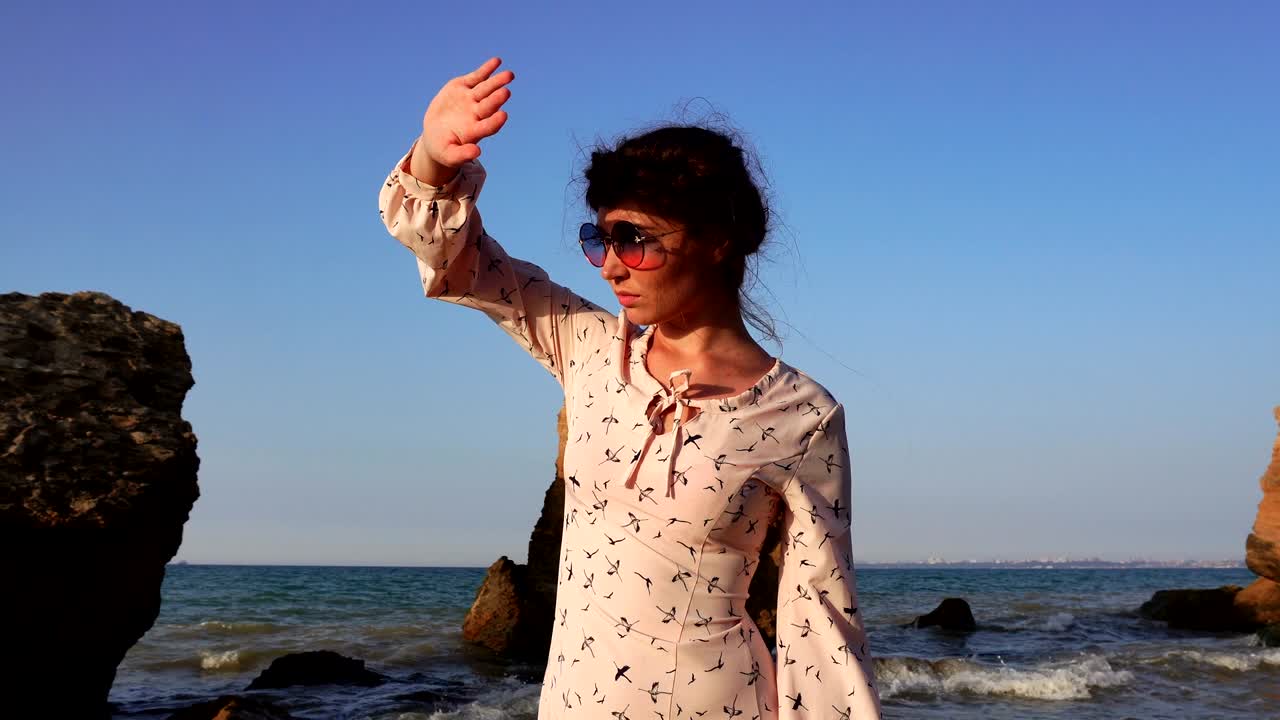 adorable chica caucásica morena con gafas de sol y vestido rosa claro se sienta en la piedra a la hora del amanecer