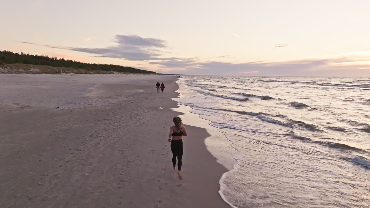 Back View Of Woman Jogging Along Serene Beach At Sunset With Waves Splashing Onto Shore. drone ascend, tracking shot