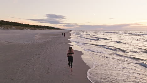 back view of woman jogging along serene beach at sunset with waves splashing onto shore. drone ascend, tracking shot