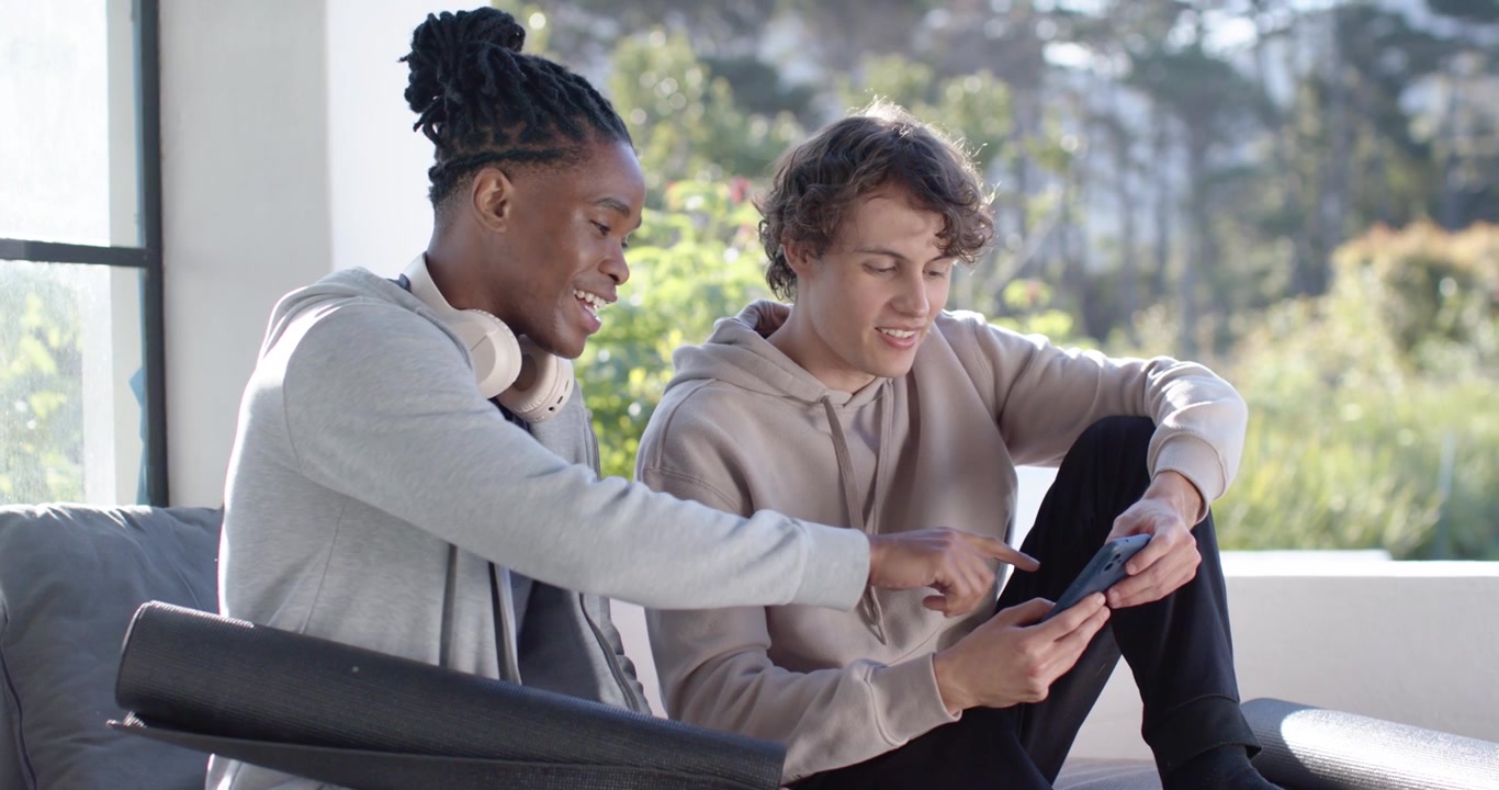 Diverse male friends at home right showing phone left pointing tapping yoga mat headphones sharing