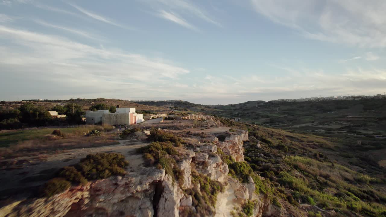 The drone flies straight toward the cliffs of Gozo, then rises to skim above them, following a winding road bathed in golden evening light
