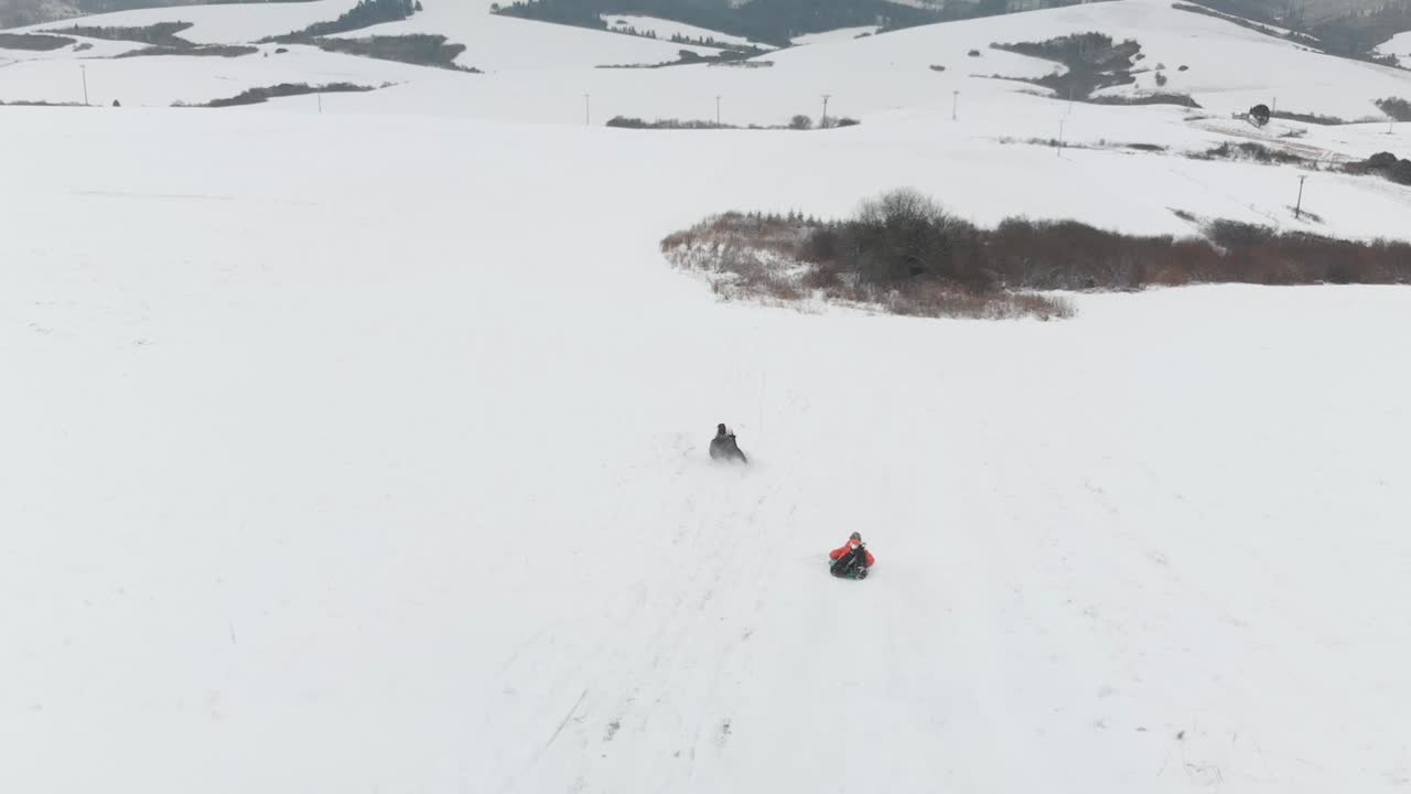 siguiente vista aérea, gente en trineo de madera bajando la colina en un paisaje de nieve invernal con el pueblo en el fondo