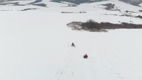 Aerial-following-view,-people-on-wooden-sleigh-riding-down-hill-in-wintery-snow-landscape-with-village-in-background