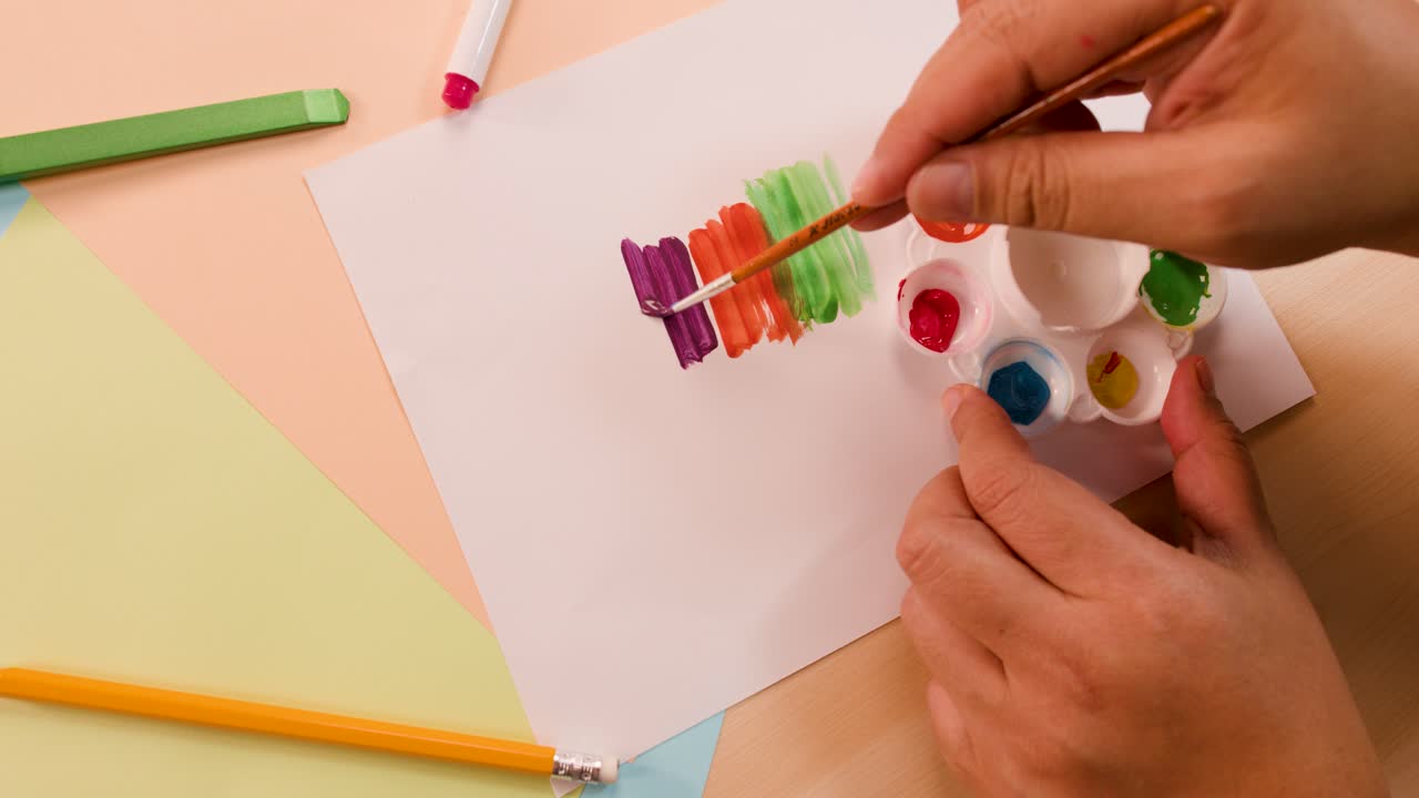 Hand uses paintbrush to create vibrant stripes on paper, surrounded by art supplies, top-down view