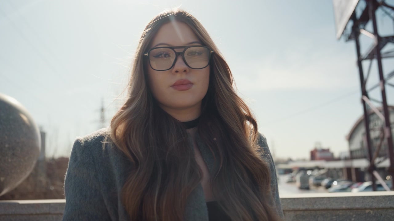 Bella donna con occhiali su una panchina di pietra, street blogger che si prepara a posare in una piazza illuminata dal sole, skyline architettonico e scultura sferica sullo sfondo, espressione rilassata, sicura e naturale.