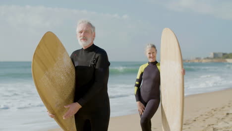 front view of a senior couple in wetsuit with surfboard standing on the sandy beach and looking at the camera