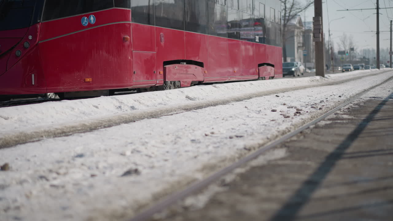 city landscape with long bus tram gliding along snowy rails between two lanes, red carriage passing storefronts and parked cars, urban public transport in motion under winter sunlight