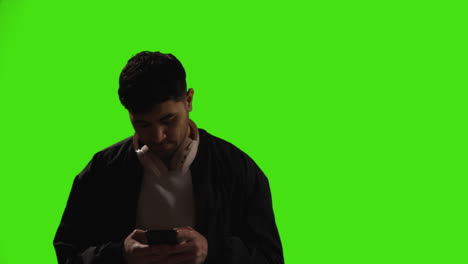 young man wearing wireless headphones text messaging on mobile phone against green screen with low key lighting