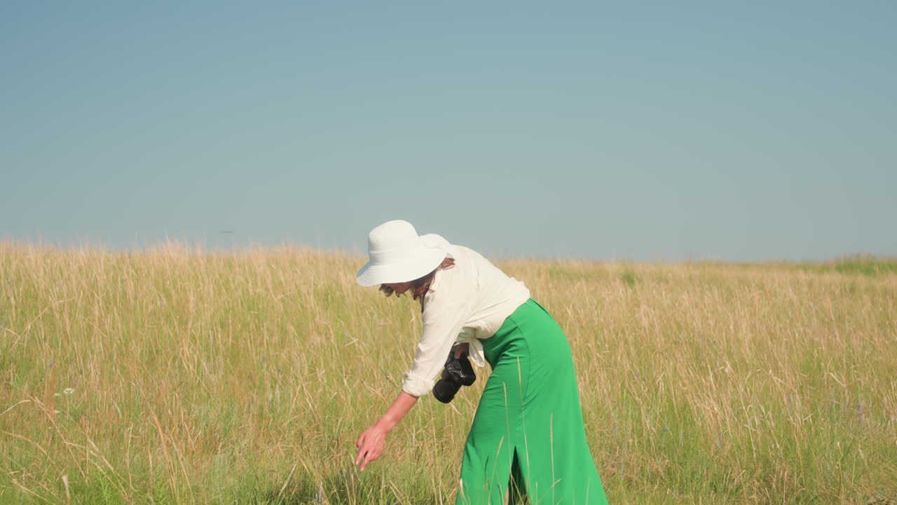 Female photographer in white blouse, green trousers, and wide hat bends forward to touch barley stalks in sunlit grassy field, holding camera in hand under clear blue summer sky