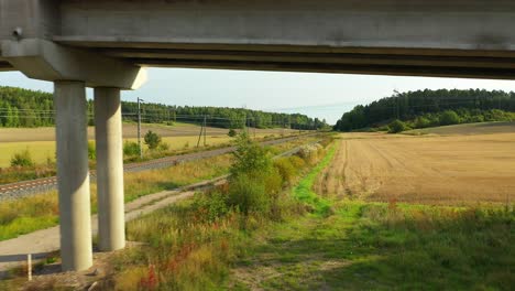 low altitude aerial drone view moving backwards following on the side of electrified railroad tracks with forests and fields on both sides, passing under road bridge