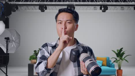 close up of asian photographer using a camera taking pictures and making shh gesture while standing in the studio