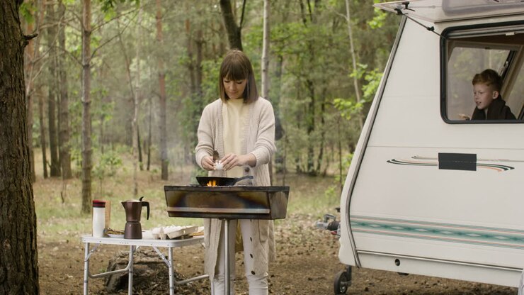 Woman cooking on the barbecue and breaking eggs in a pan while her son looking out of campervan window