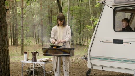 그녀의 아들이 campervan 창 밖을 내다보는 동안 바비큐 요리를 하고 냄비에 달걀을 깨는 여자