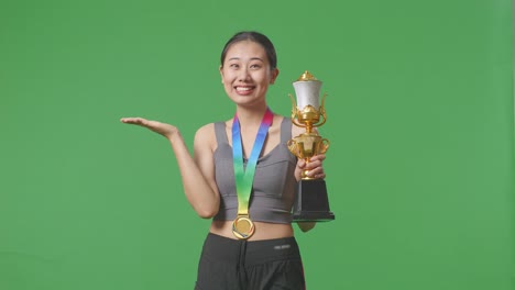 asian woman with a gold medal looking at a gold trophy in her hands, smiling, and pointing to side on green screen background in the studio