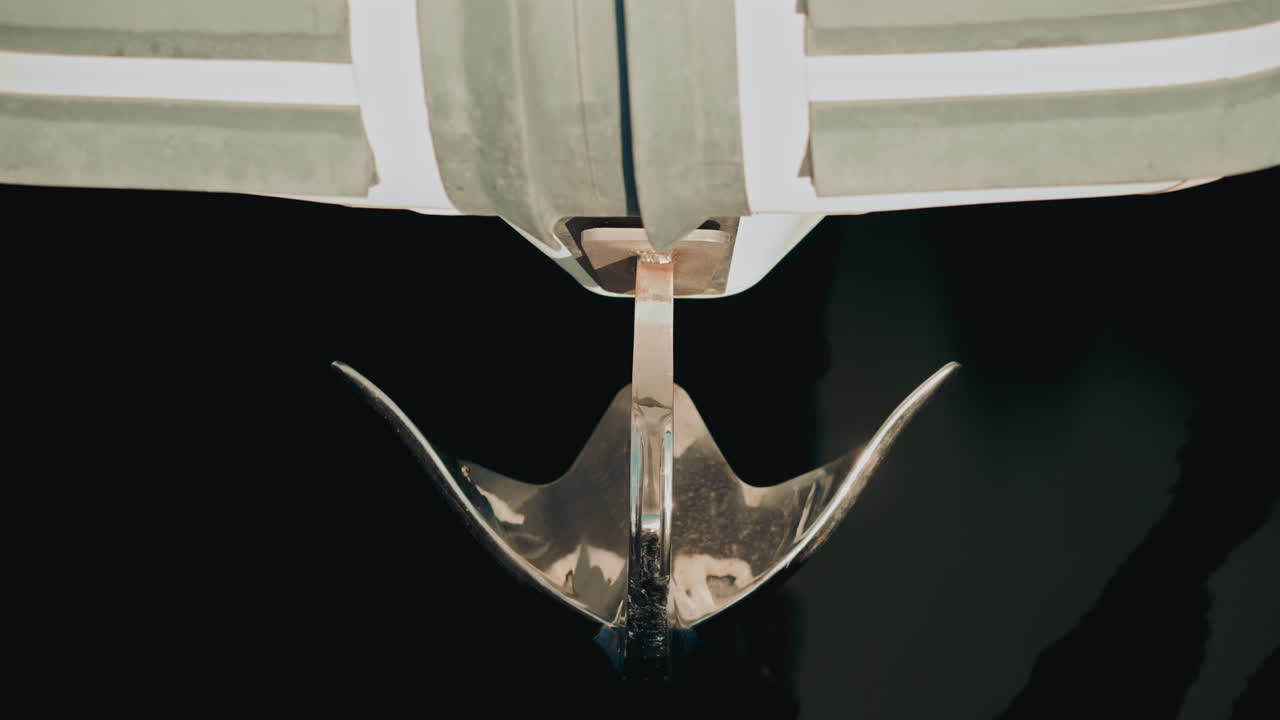 Close up of a polished stainless steel yacht anchor hanging above calm dark water, reflecting soft daylight and marine surroundings