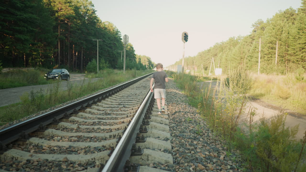 rear view of kid in casual wear walking carefully along rail track surrounded by gravel and vegetation, with background featuring car on nearby forest road, power poles, signal light, and tall trees