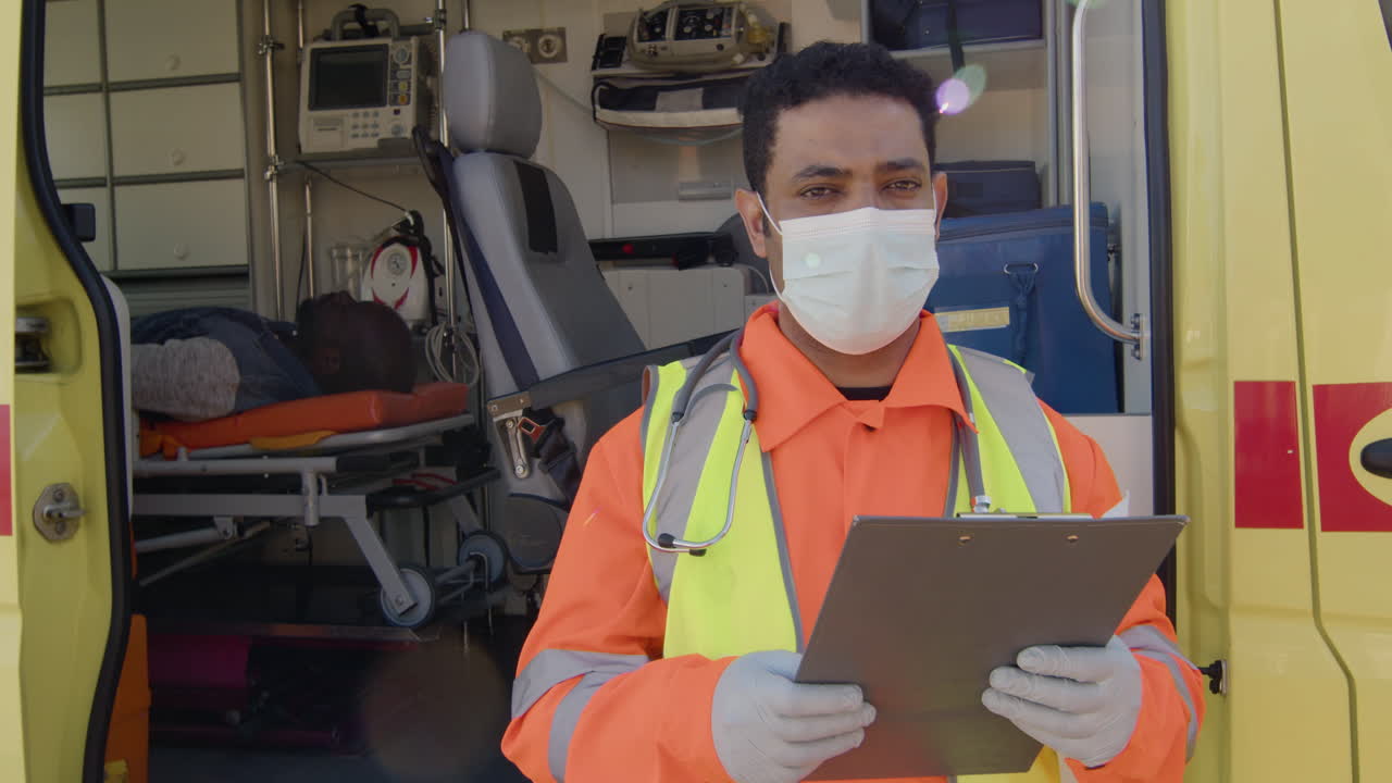 retrato de un paramédico latino con mascarilla parado frente a una ambulancia, sosteniendo un portapapeles y mirando la cámara