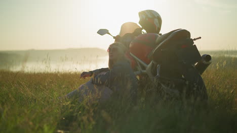 a woman wearing a flower headband and sunglasses sits peacefully in a grassy field, leaning back against her motorcycle as the sun sets in the background