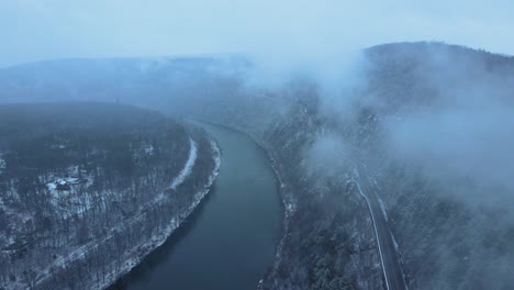 Aerial-footage-of-a-snowy,-scenic-byway,-winding-mountain-valley-road-during-a-snowstorm-with-pine-trees,-a-river,-mountain-highway,-rocky-cliffs,-and-forests-during-winter-on-a-cold,-blue-day