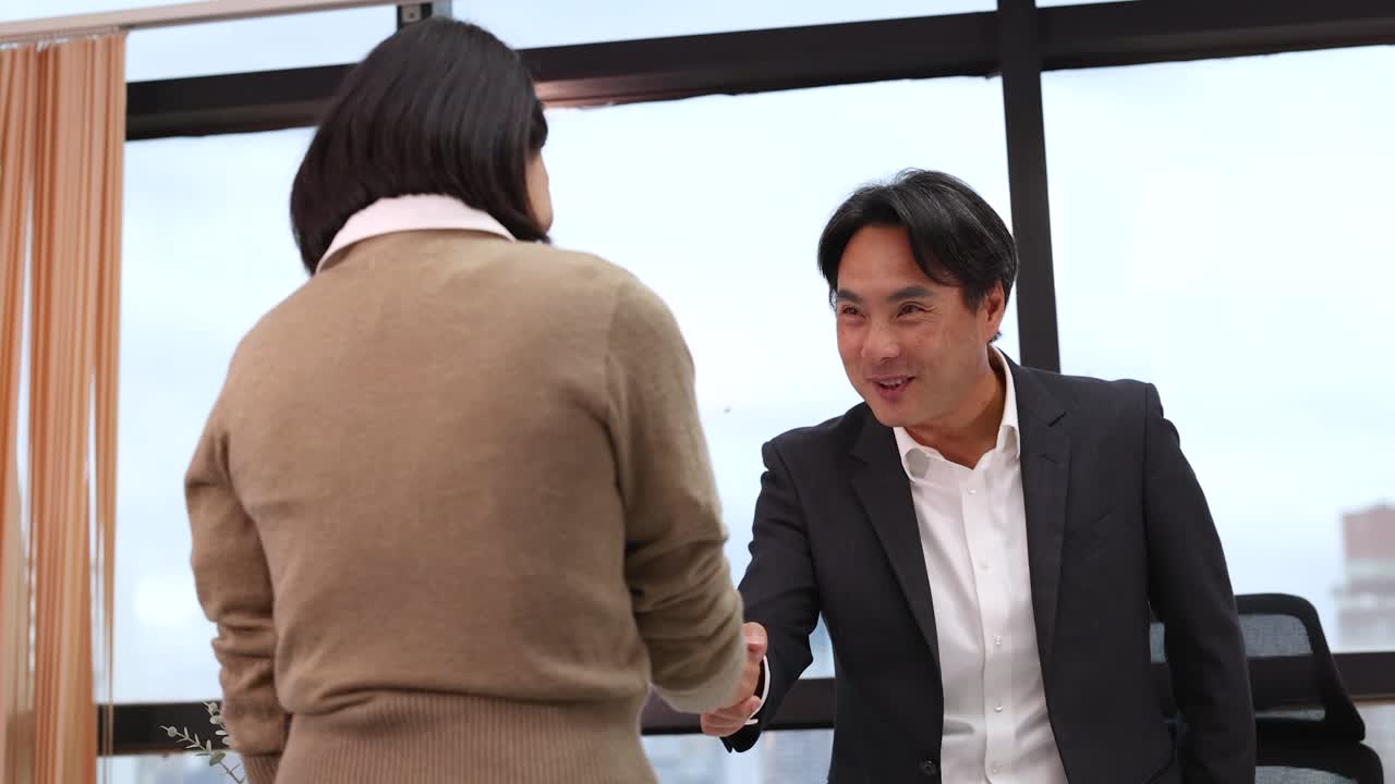 明るい企業のオフィスで、アジア系のプロフェッショナルな男女が机を挟んで握手をしている。