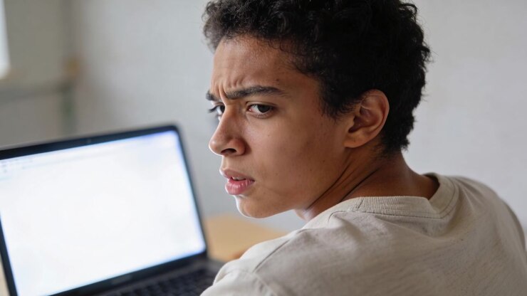 Turning teen in light tee reacting to noise at home desk, processing, refocusing on laptop