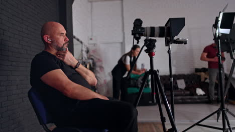 calm caucasian man wearing black clothes sits on the chair near the camera. man and woman at backdrop recording video in studio.