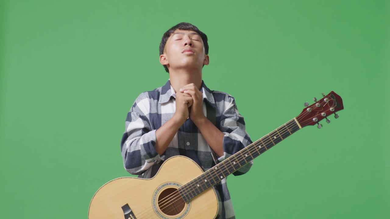 기타를 들고 있는 아시아 십대가 녹색 스크린 배경에서 무언가를 위해 기도하고 있습니다.