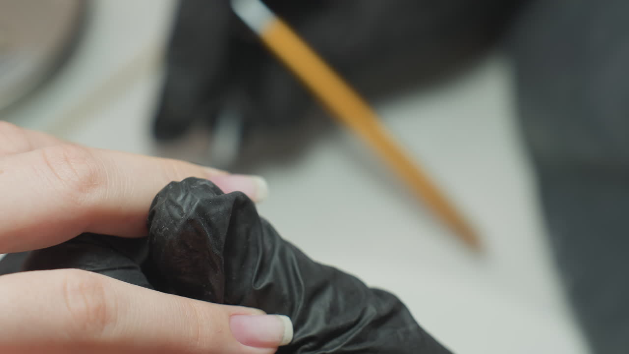 Close-up of nail technician wearing dusty black glove delicately using nail polish brush to apply polish to client's fingernail with precision during professional manicure in clean salon environment