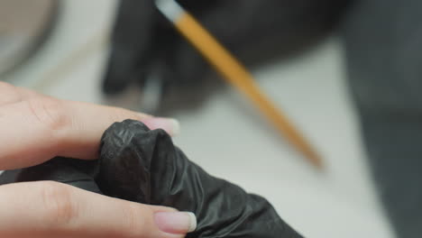 close-up of nail technician wearing dusty black glove delicately using nail polish brush to apply polish to client's fingernail with precision during professional manicure in clean salon environment